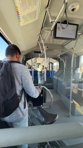 The video shows the interior of a bus with a man wearing a backpack standing near the front of the bus. A bicycle is visible next to him. The bus appears to be in motion as the view slightly shifts. The interior of the bus has rows of blue seats and overhead handrails. A screen is mounted on the ceiling displaying some information and a crest. The man is holding onto a metal pole. The lighting suggests it is daytime.