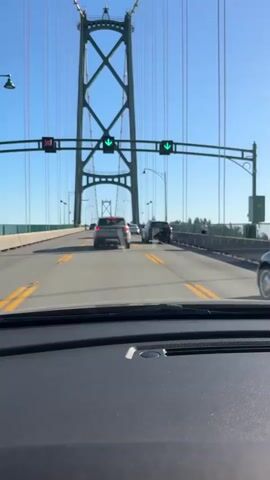 The video is filmed from the perspective of a driver, looking forward through a car's windshield. The car is on a multi-lane bridge, with traffic moving forward. A large green suspension bridge tower with a lattice structure dominates the upper portion of the frame. Traffic lights are visible above the lanes, with two showing green arrows pointing downwards, indicating traffic can proceed. Several cars are on the bridge, including a silver SUV directly ahead, a blue Volkswagen Beetle to the right, and other vehicles further ahead and to the left. The road has yellow lane markings. The sky is clear and blue. The overall environment appears to be a sunny day.