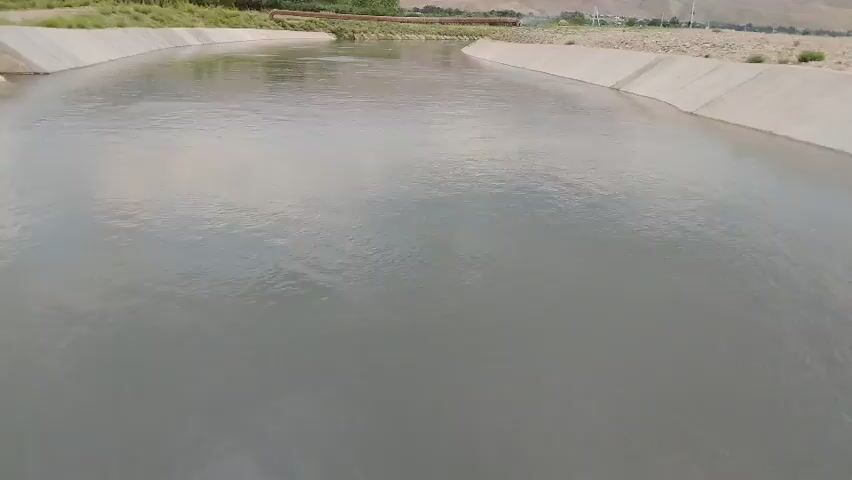 The video shows a wide, greyish river flowing through a concrete canal. The canal has a curved shape, with concrete walls on either side. The water appears calm, with only a slight ripple on the surface. The banks of the canal are lined with dry, sandy soil and some sparse vegetation. In the distance, there are some buildings and hills, suggesting a rural or semi-arid environment. The sky is overcast, as indicated by the diffused light and the greyish tone of the water. There is no visible text in the video. The scene conveys a sense of a functional waterway, possibly for irrigation or transportation, in a natural, unpopulated setting. The time of day appears to be daytime, given the ambient light.