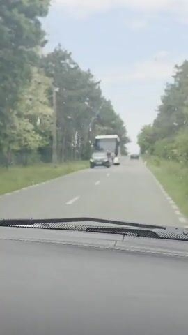 The video shows a car driving on a road flanked by trees.  In the distance, a bus is stopped in the road.  A police car is parked in front of the bus, and people are standing near the bus.  The car filming passes by, and the road continues to stretch onward into the distance.