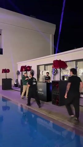 A night scene outdoors by a swimming pool. Several people are near a DJ booth with speakers and lights. One person, wearing a dark shirt, is walking away from the DJ booth towards the left. Another person, in a green hoodie and khaki pants, is standing in front of the DJ booth, holding a phone to take a picture or video. Two people are behind the DJ booth, one appearing to be a DJ, and another is standing beside them. The DJ booth has logos that read "GOLD LEAP" and "WINWIN". Red and white speakers are positioned on stands to the left and right of the DJ booth. The swimming pool has blue water and is in the foreground.
