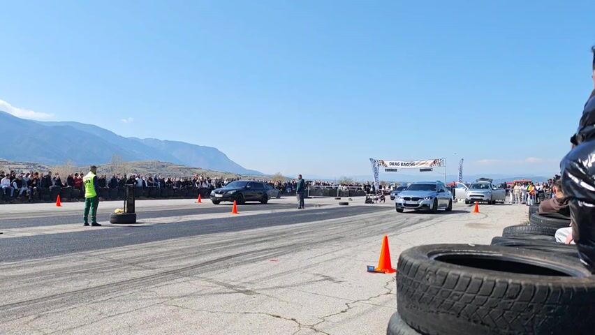 The video shows a drag race between a silver BMW sedan and a dark-colored station wagon. The BMW does a burnout and then takes off down the track ahead of the station wagon. There is a large crowd of spectators watching the race from the side of the track. The track is located in a rural area with mountains in the background.