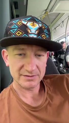 The video shows a man wearing a brown t-shirt and a baseball cap sitting on a bus. He is looking at the camera and smiling. In the background, other passengers are visible. The interior of the bus is dimly lit.