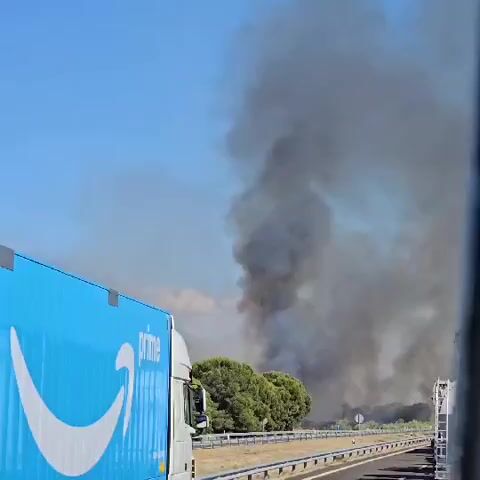 The video captures a scene on a highway where a large plume of dark smoke billows into the sky, indicating a fire. An Amazon Prime truck, with its distinctive blue and white branding, is prominently featured in the foreground on the left side of the screen. Other trucks are also visible on the highway, suggesting a traffic slowdown or blockage due to the fire. The environment appears to be sunny with a clear blue sky, contrasted by the dense smoke. There are trees and roadside barriers present. No people are clearly visible in the footage.