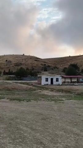 The video shows a wide shot of a rural landscape under a cloudy sky. In the foreground, there is a dirt path. In the middle ground, there are a few small, simple buildings. One building is white with a dark door and windows, and it appears to be made of brick or stone. To its right, there is a structure with a red roof providing shade over an outdoor seating area. Further in the background, a hill rises with sparse vegetation and a few scattered trees. The sun is setting or has recently set behind the crest of the hill, casting a bright, hazy glow that creates a halo effect in the sky. There are a few indistinct objects or vehicles visible on the distant hilltop. The overall impression is one of a quiet, somewhat desolate rural setting at dusk.
