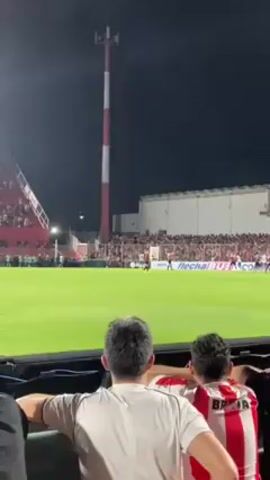 From an elevated position, the video shows an outdoor night soccer match taking place in a stadium. The field is well-lit. The camera focuses on the backs of three spectators wearing team colors, with their gazes fixed on the game. The stands are packed with fans. Red and white vertical stripes are prevalent in the stadium's design, including a tall communications tower and the stands themselves. The sky is dark. The scene suggests a tense or exciting moment in the match.