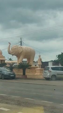 The video begins showing a large, light-colored elephant statue with intricate carvings on its side. To the left of the elephant is a building with a dome and ornate pillars. To the right of the elephant is a statue of a man in traditional attire holding a spear. In the foreground, a dark gray car is driving past the elephant statue. The video then shifts to show a white car driving past the same elephant statue. The background consists of a cloudy sky and power lines. The environment appears to be a roadside with a paved surface and some vegetation. The word "DEVAGANDHA" appears briefly on screen during the video.