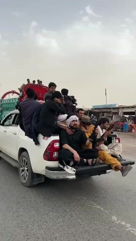 Several men are riding in the back of a white pickup truck on an asphalt road. The men are wearing traditional clothing, and some have turbans. Several are holding rifles. Other men on motorcycles travel near the pickup truck. A brightly-colored truck with elaborate decoration drives in the background. The sky is overcast. Buildings are visible on the side of the road.