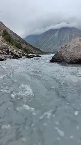 A low-angle, handheld video captures a churning river flowing through a mountainous landscape. The water in the foreground is choppy and gray with white foam. To the left, a rocky bank is visible, with a few small trees growing on it. Beyond the river, rugged mountains rise steeply, their peaks obscured by heavy, gray clouds. The sky is overcast.