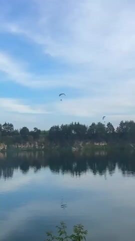 This is a vertical, outdoor shot of a body of water with a tree-lined cliff face and a blue sky with some clouds. There are two paragliders in the air. One paraglider, with a person visible beneath it, is in the upper left quadrant of the frame. The other paraglider is smaller and further to the right. The water reflects the sky and the trees. The lighting suggests it is daytime.