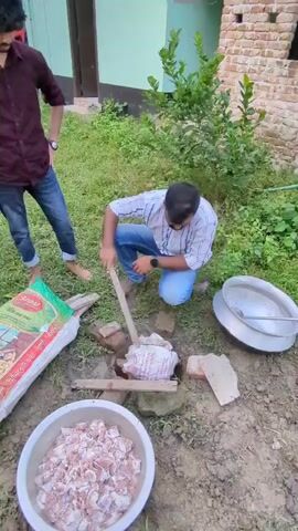 The video shows an outdoor scene with several individuals. In the foreground, two men are tending to a cooking setup over an open fire. One man, wearing a maroon shirt and jeans, stands observing, while another man, in a checkered shirt and jeans, kneels and uses a stick to stir something in a pot. Next to them is a large, colorful bag and a bowl filled with raw meat. In the background, there are trees, a building, and more people interacting. The setting appears to be a rural or semi-rural environment, possibly a gathering or event.