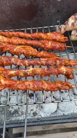 A close-up shot captures several skewers of twisted meat grilling over charcoal. The meat appears to be coated in a reddish marinade and is developing a char in places. The grill grate is dark and speckled with ash. In the upper right corner, a piece of what looks like grilled chicken leg is visible, indicating a variety of items are being cooked. The focus is on the meat as it cooks, with the camera slowly panning across the skewers.