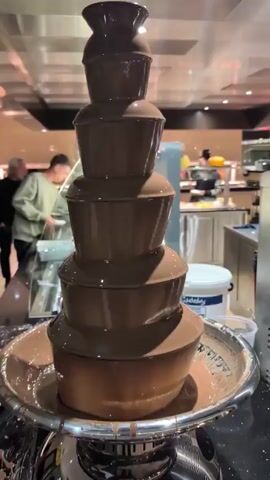 The video begins with a close-up of a chocolate fountain, the chocolate smoothly cascading down its tiers. The camera then pans right, revealing a stainless steel ice cream machine. Continuing to pan right, the scene widens to showcase a dessert buffet station. The area is well-lit with stainless steel surfaces and a few people in the background browsing the food options.