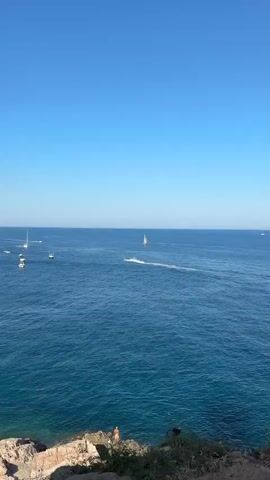 The video opens with a wide shot of a vast expanse of deep blue ocean under a clear, bright blue sky. In the distance, a solitary white sailboat with two sails is visible. Closer to the camera, a jet ski is rapidly moving across the water, leaving a white spray trail behind it. The camera then pans slightly to the left. The jet ski continues its movement, and the sailboat remains in the distance. The video transitions to a wider view of a bay filled with numerous boats anchored in the turquoise water. The shoreline is rocky and covered in vegetation. On the rocky shore, a few people can be seen standing or sitting. The horizon shows more sailboats scattered across the water, and a small island is visible in the bay. The sky remains clear and blue throughout.