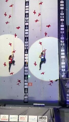 Two female athletes are rock climbing on an indoor climbing wall. Both athletes are wearing harnesses and climbing shoes. They are ascending the wall, with one athlete on the left side and the other on the right. The wall has numerous red climbing holds shaped like stars. There are two large white circles of light illuminating portions of the wall where the athletes are climbing. A digital timer is visible at the top of the wall, displaying the athletes' progress. The overall environment appears to be an indoor sports arena or stadium.