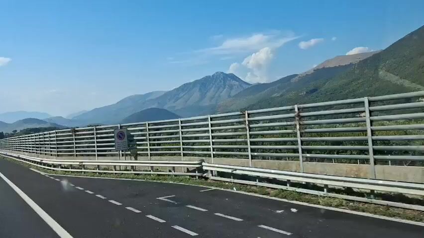 A car is driving on a highway with mountains in the background. The highway has multiple lanes and a sound barrier along the side. The sky is blue with some clouds. There are other cars on the highway.