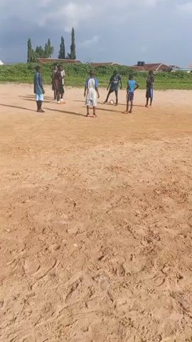 A group of boys are playing soccer on a dirt field. They are wearing casual clothing. The field is dry and sandy with some sparse grass in the background. The sky is cloudy.