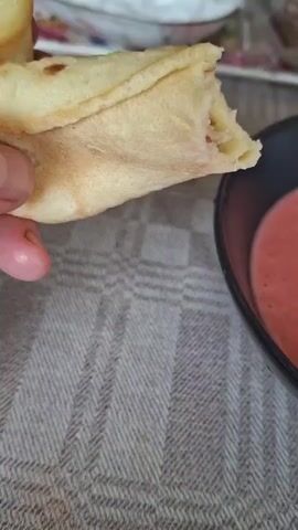 A hand holds a piece of flatbread or tortilla and dips it into a bowl of pinkish-red, creamy dip. The flatbread is folded and appears to have a filling. The bowl is black and sits on a textured, woven surface, likely a tablecloth. The lighting suggests an indoor setting, possibly a mealtime.