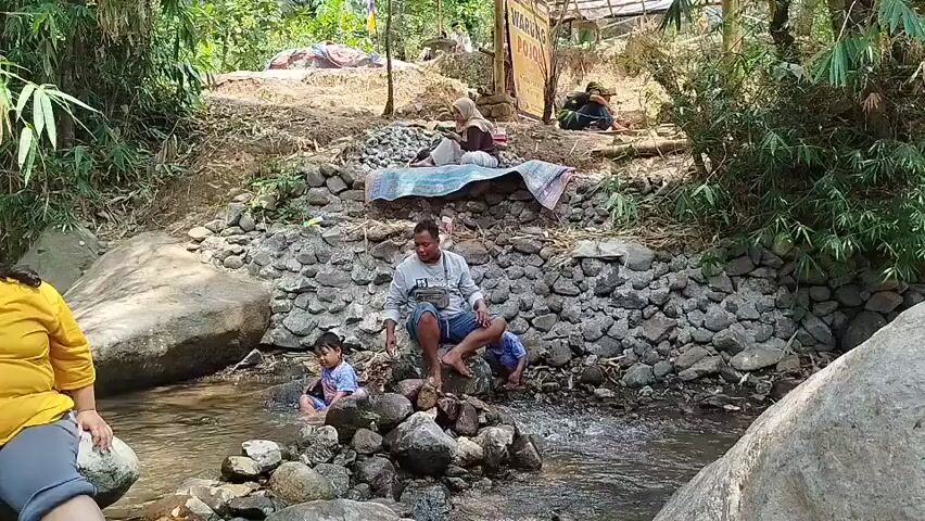 The video shows a natural environment with a stream flowing through it. In the foreground, a person wearing a yellow shirt is seated on a large rock to the left. A man is seated on rocks in the middle of the stream, with two children nearby. One child is in the water and another is partially submerged. In the background, two people are sitting on a mat on a raised area. There are rocks, trees, and bamboo plants surrounding the stream. A sign is visible in the background. The overall scene depicts a relaxed outdoor setting, possibly a recreational area near a stream.
