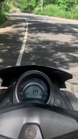 The video begins with a close-up of a motorcycle's dashboard, showing a speed of 27 km/h. The view then pans to the right, revealing a winding road bordered by lush green vegetation on both sides. In the distance, a waterfall cascades down a rocky cliff face. Several vehicles, including cars and what appear to be tuk-tuks, are visible on the road. The sun is shining, casting dappled shadows on the road surface. The overall scene depicts a scenic drive through a mountainous, tropical landscape.