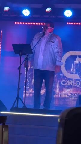 A man stands on a stage wearing a light-colored button-down shirt over a white t-shirt and blue jeans. He is holding a microphone and appears to be speaking or singing. There is a music stand to his left. The stage is lit with blue and pink lights, and a large screen behind him displays a cityscape and the letters "GC" and "CARLOS". The overall lighting is dim and atmospheric, suggesting a performance or event setting.
