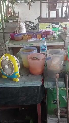 The video shows an outdoor market stall. In the foreground, a yellow fan with a cartoon face sits on a blue surface. Behind the fan are several plastic containers and a bottle of water. In the background, a young man in a black t-shirt stands at a counter, and two women are also visible. The stall is made of wood and metal, and there are various items displayed, including bags of food and bowls. A motorcycle is parked behind the stall. The overall environment appears to be a market setting, possibly in a Southeast Asian country given the visual cues.