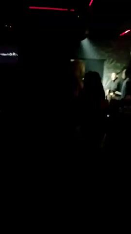 The video is shot in a dark and crowded nightclub. The lighting is colorful and appears to be coming from stage lights. Initially, a man is seen touching a woman's buttocks. He is wearing a dark shirt, camouflage shorts, and white sneakers. The woman is wearing a bikini-style outfit and bright pink platform heels. They are standing near a small round table with drinks and a partially visible person sitting. Later, the camera pans around the room showing other people milling about near the table.
