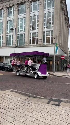 The video shows a group of people riding on a multi-person pedal-powered vehicle, referred to as a "Beer Bike," on a city street. The vehicle is decorated with purple accents and has the words "BELFAST Beer Bike" visible. Several women are seated on the sides of the bike, pedaling, while one person stands at the back, possibly steering or serving. Another person is standing on the back of the bike. In the background, there is a large building with multiple floors and windows, with storefronts visible at street level, including "DUNNES STORES." A black car is parked on the left side of the street, and other pedestrians are visible further down the sidewalk. The scene appears to be a casual, recreational outing in an urban environment.