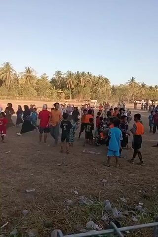 A wide shot shows a group of people, mostly children, gathered on a dry, dirt field under a clear sky. Trees line the horizon in the background. Many children are wearing athletic clothing, suggesting a sporting event or practice. Some are standing, while others appear to be in motion. The atmosphere seems to be casual and possibly energetic, with individuals interacting and moving around the field. The ground is uneven and shows signs of wear.