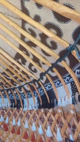 The video provides an interior view of a yurt, a traditional nomadic dwelling. The camera pans across the structure, revealing wooden poles forming the roof and walls. Decorative textiles are visible, including a patterned fabric with spiral motifs covering the upper portion of the interior and a fringe of tassels along the lower walls. The overall impression is one of a rustic and culturally rich living space.