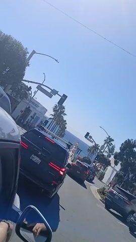 The video shows a street scene, filmed from inside a car moving through traffic. Palm trees and building facades line the street. In the distance, the ocean horizon is visible. Traffic lights appear green. Several vehicles, including SUVs, are visible in the frame. The sky is clear and blue. A side mirror of the car filming the scene is visible, showing a partial reflection of the interior.