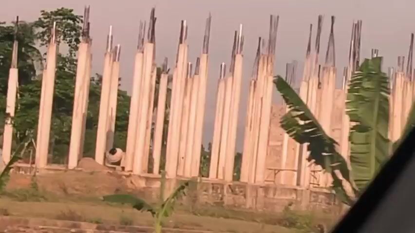 The video shows a building under construction. The building consists of many tall, white columns with steel rebar extending from the top. There is a small, red-roofed building next to the construction site. Green trees and other plants are visible in the background. Cars drive by on a road in front of the construction.