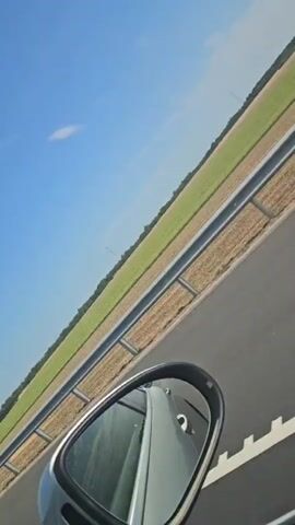 The video shows a view from inside a car, looking out the passenger side window and at the side mirror. The car is on a paved road, likely a highway, with a metal guardrail to the right. Beyond the guardrail are fields of green and brown crops stretching to the horizon under a clear blue sky with a few scattered clouds. The side mirror reflects the interior of the car, including a seat. The camera is angled, giving the impression of movement. There are no people visible in the frame.