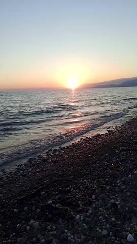 The video shows a sunrise over a rocky beach and the ocean. The sun is partially obscured by clouds on the horizon, casting a warm glow across the sky and water. Gentle waves lap at the shore, which is covered in smooth, dark pebbles. The overall impression is one of peace and natural beauty.