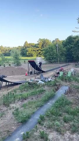 A person on a dirt bike is performing a jump. The dirt bike is airborne, suspended against a clear blue sky. Below the bike, ramps and dirt mounds are visible, suggesting a motocross track or stunt setup. In the background, trees and foliage frame the scene, indicating an outdoor environment. The video transitions to show the dirt bike rider approaching a large inflatable airbag, labeled "DD AIRBAGS." The rider then launches off a ramp and lands on the airbag, completing the stunt. The environment is rural or semi-rural, with evidence of recreational activities like a pool and trampoline also present in the background.