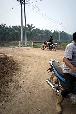 A dirt road passes through a palm oil plantation. In the foreground, a person is seated on a motorcycle. In the middle ground, another person is riding a motorcycle away from the camera, crossing a small bridge. Power lines and poles are visible in the background. The sky is overcast.