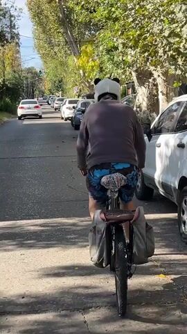The video shows a person riding a bicycle on a street lined with parked cars. The person is wearing a gray sweatshirt, blue shorts with a floral pattern, and a white panda hat. The bicycle is equipped with two saddlebags. The road is paved, and the weather appears sunny. The camera is positioned behind the cyclist, initially capturing a view of their back and then transitioning to a close-up of the bicycle handlebars. The bicycle handlebars have stickers of “CANNA BISS” covering the pole that connects the bars to the front wheel. As the clip continues, the camera returns to the rear perspective of the cyclist.