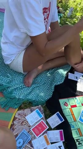 The video captures an overhead view of a Monopoly game being played outdoors on a blanket spread over grass. Three individuals' legs and feet are visible, indicating they are seated around the board. The game board is bright green with colorful property squares and various game cards are scattered on the blanket, along with Monopoly money. Two dice have been rolled and are on the board, and one player's hand is reaching towards them, suggesting an active turn. The scene is bathed in sunlight, with green foliage visible in the background, suggesting a park or backyard setting. The overall atmosphere appears casual and relaxed, as if friends are enjoying a game day.
