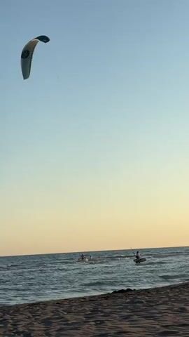 A wide shot of a beach with calm ocean waves lapping at the shore. The sky is clear with a gradient of blue to yellow. In the distance, on the water, are several figures. To the right, a person on a paddleboard is visible. To the left and further out, two more figures are in the water. High in the sky, a kiteboarder is in action, their kite clearly visible. The sand in the foreground is textured and the water has small waves.