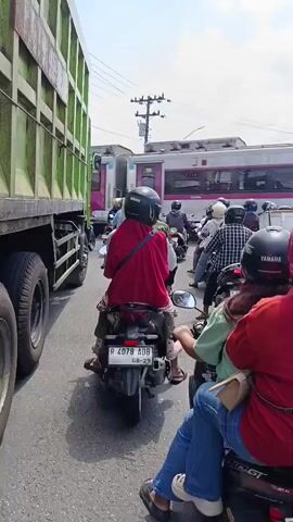 A traffic jam occurs on a street with multiple lanes of vehicles, including motorcycles and a large green truck. In the background, a train with "KAI" branding is visible, with some of its cars appearing to be detached. The sky is cloudy.