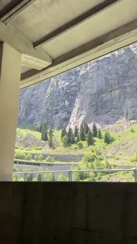 The video shows a view through a concrete structure. The view is of a steep, rocky mountain face with patches of green vegetation and trees. There are also visible remnants of what appear to be old structures or roads on the mountainside. The camera is panning slowly to the right.