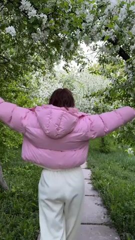 A woman is walking away from the camera on a narrow pathway made of square concrete slabs. The woman is wearing a light pink puffy jacket, white wide-legged pants, and white sneakers. The camera angle is low, shooting up towards the woman. She raises her arms out to her sides and over her head. The path is surrounded by green grass and flowering trees. The trees are full of white blossoms, creating a canopy over the path. The trees are full of green leaves. The light in the video is diffused. The background is blurred due to the depth of field.