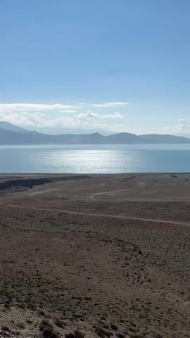 The video presents a panoramic view of a vast, arid landscape under a clear, bright blue sky. A large, light blue lake dominates the midground, its surface shimmering intensely with reflections of bright sunlight. In the foreground, the terrain is dry, rocky, and sparsely vegetated in shades of brown and tan, with a dirt track winding across it. A solitary white utility pole is briefly visible on the left side of the frame at the beginning.

The camera executes a slow, smooth horizontal pan to the right, gradually revealing the expansive vista. Distant mountain ranges line the horizon beyond the lake. As the camera pans further right, a series of small, distant wind turbines become visible across a flat expanse beyond the lake, followed by more distinct snow-capped peaks in the background. Scattered white clouds drift across the otherwise clear sky.

The scene conveys a sense of serene vastness and natural beauty, characteristic of a high-altitude region like Gyirong County, China. The time of day appears to be mid-morning or early afternoon, with clear, sunny weather. No people or specific activities are depicted within the frame, emphasizing the remote and untouched quality of the environment. There is no visible text.