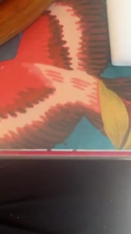 The video shows a close-up shot of a sizzling steak dish served with fried potatoes. The steak is cut into thick slices, still rare, and sprinkled with coarse salt. The potatoes are golden brown and arranged around the meat on a hot plate. The plate sits on top of a wooden tray with a colorful bird design. Background sounds include indistinct voices and upbeat music.