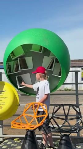 A young boy stands in front of two green, spherical structures that serve as outdoor bookshelves. He is wearing a light gray t-shirt, dark gray shorts, and a red baseball cap. He is holding a book.  Around him are several brightly colored, modern-looking chairs. The boy is near a fence. Behind the fence is a building with a white facade. The day is sunny with a clear blue sky. The boy looks from one bookshelf to the other and then looks at the book he is holding.