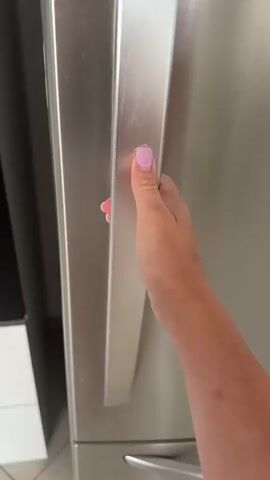 The video opens with a close-up of a hand with painted nails reaching into a refrigerator. The hand grasps the handle of a drawer, which is then pulled open to reveal the contents of the refrigerator. Inside the refrigerator, on the top shelf, is a wooden bowl containing plastic toy food items: a strawberry, a lemon, a slice of onion, and what appears to be a piece of chocolate or a toy steak. A can of Stella Artois beer and a slice of watermelon are also visible on the top shelf. The drawer below contains various vegetables, including red peppers and green cucumbers. Another drawer beneath that holds a dark bottle with a yellow cap and a package of cigarettes. The camera then zooms in on the wooden bowl and its contents. The scene appears to be indoors, in a kitchen environment, with the refrigerator door open.
