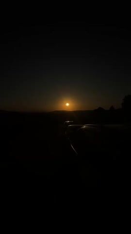 A video captures a sunset with a partial solar eclipse. The sun, partially obscured by the moon, appears as a crescent shape against a dusky sky. The foreground is dark, with the silhouettes of cars visible. The sun's light creates lens flares in the image.
