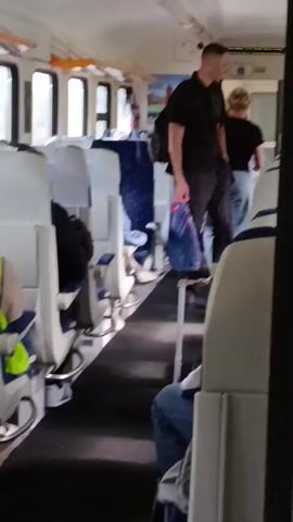 A man in a black polo shirt and dark pants walks down the aisle of a train car. He carries a blue plastic bag in his right hand. The train car has rows of white and blue seats. The man is moving away from the camera.