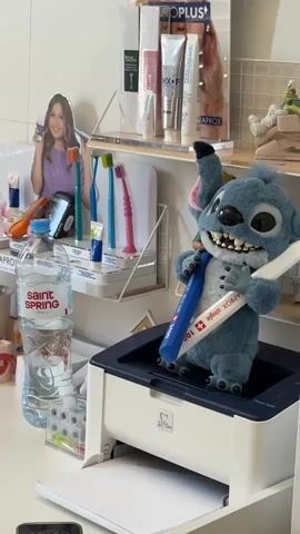 A blue plush Stitch toy stands on top of a white printer, holding a blue and white object in its hands. Behind the Stitch toy, on a shelf, are various dental hygiene products, including toothbrushes and tubes of toothpaste. To the right of the printer and Stitch is a glass-fronted cabinet filled with more dental hygiene products, predominantly small boxes with the brand name "CURAPROX" visible. The lighting is bright, suggesting an indoor environment, likely a dental office or a retail display. The background shows more shelves with products and a poster of a young woman.
