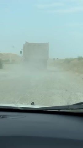 The video begins by showing the rear of a dump truck driving away on a dirt road, kicking up a large cloud of dust. The camera is positioned from inside a vehicle, looking through the windshield. In the distance, another truck can be seen. The environment appears to be dry and rural, with sparse vegetation and rolling hills in the background. The sky is hazy. The footage continues with the dump truck moving further down the road, still enveloped in dust, and the camera remains stationary.